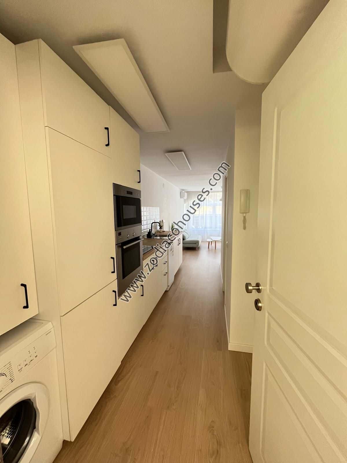 La imagen muestra una cocina estrecha, de estilo galera, con gabinetes blancos y pisos de madera clara. En el lado izquierdo, hay una lavadora blanca debajo del mostrador, con un horno y un microondas integrados encima. El lado derecho presenta una puerta blanca con un pomo plateado, que conduce a otra habitación. En el fondo, se puede ver un vistazo a una sala de estar o comedor con una pared blanca y un piso de color claro. La cocina parece estar bien iluminada, con luz natural entrando por la puerta abierta a la derecha. El diseño general es moderno y minimalista, con una apariencia limpia y ordenada.