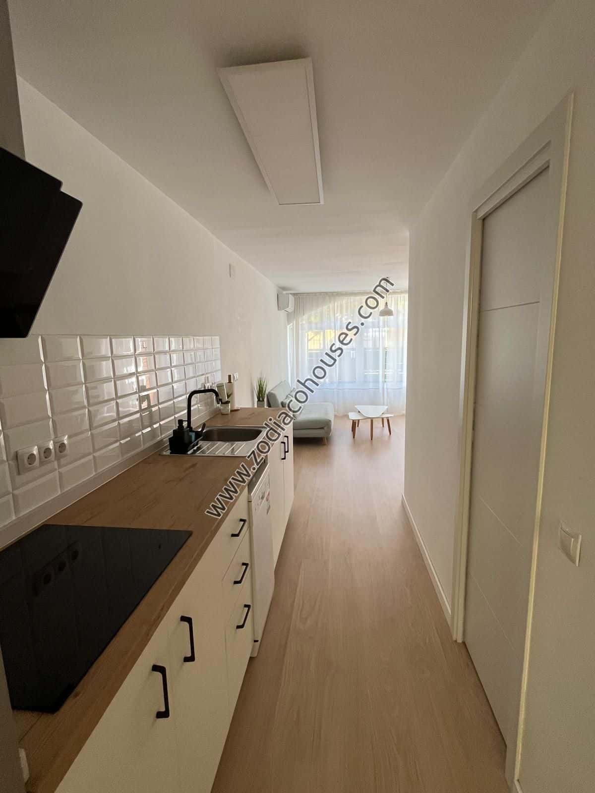 Una cocina moderna con armarios blancos y un respaldo de ladrillo blanco. La encimera es de madera, y hay un fregadero con un grifo plateado. A la derecha de la cocina, hay un pequeño comedor con una mesa blanca y sillas. El suelo es de madera clara, y las paredes son blancas. Hay una puerta al final de la habitación, y una ventana sobre el fregadero deja entrar la luz natural.